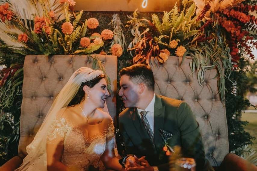 Novios sentados en sus sillas con una decoración floral detrás